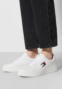 Zapatillas de cuero blancas con punta redondeada, diseño perforado y marca prominente en el lateral. Combinadas con pantalones de mezclilla negros.