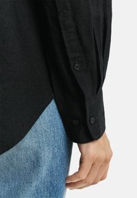 Zwarte linnen blouse met lange mouwen en knoopmanchetten, met een afgeronde zoom. Gecombineerd met lichtblauwe spijkerbroeken, die het textuurcontrast benadrukken.