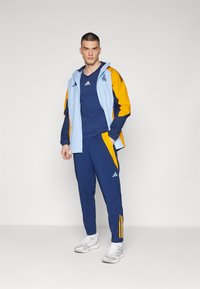 Adidas trening odijelo koje se sastoji od svijetloplave i žute jakne, tamnoplavog gornjeg dijela i odgovarajućih hlača s žutim detaljima te bijelih tenisica.