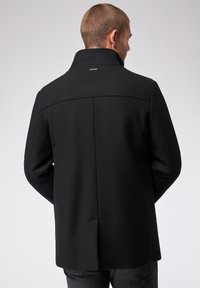 Zwarte wollen jas met een hoge kraag, strakke lijnen en een achtersplit. Voorzien van een subtiel merkteken bij de nek. Soepele textuur, getailleerde pasvorm.