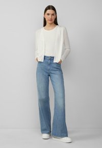 Weiße, gestrickte Strickjacke über einem weißen Top, kombiniert mit hellblauen Schlagjeans und weißen Sneakers. Weiche Texturen und lockere Passform insgesamt.