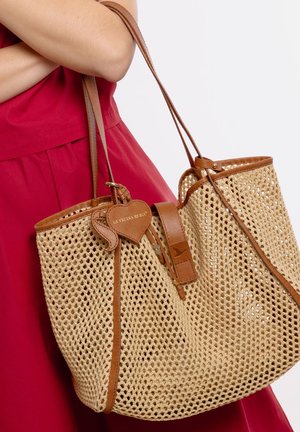 Persona con vestido rojo sosteniendo un gran bolso beige tejido con ribete y correas de cuero marrón, con una etiqueta en forma de corazón.