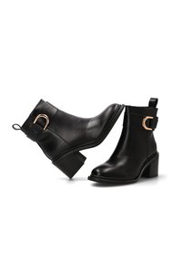 Botas tobilleras de cuero negro con tacones de bloque, que presentan un detalle de hebilla metálica y suela texturada. Punta redondeada con una lengüeta en la parte posterior.