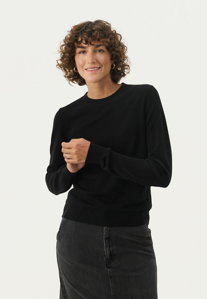 Schwarzer Strickpullover mit langen Ärmeln und gerippten Bündchen, mit rundem Halsausschnitt und tailliertem Schnitt. Getragen mit einem dunklen Rock.