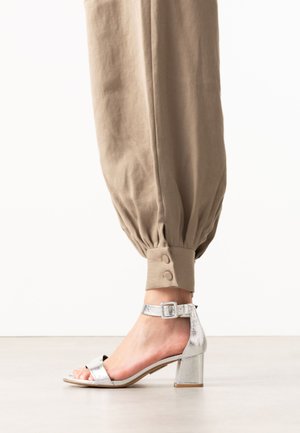 Voet met zilveren sandaal met blokhak en enkelbandje, gecombineerd met beige taps toelopende broek met knoopmanchetten.