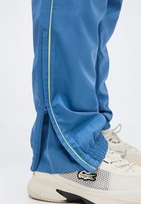 Κάτω μέρος ποδιού που φοράει μπλε φόρμα με πλευρικό φερμουάρ και νεον κίτρινη ρίγα, συνδυασμένο με λευκά και γκρι αθλητικά παπούτσια με μαύρο λογότυπο κροκόδειλου.