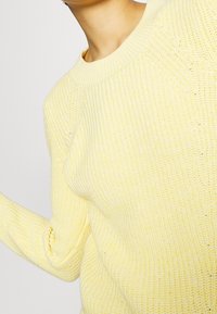 Ljusgul stickad tröja med rund halsringning och långa ärmar med ribbat mönster. Tyget verkar mjukt och något löst sittande.