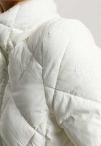 Chaqueta acolchada blanca con cuello alto, con costuras tonales y un parche de logo redondo en el hombro. Textura suave y lisa.