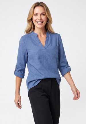 Lachende vrouw met schouderlang blond haar, gekleed in een blauw hemd met lange mouwen en een zwarte broek tegen een effen achtergrond.