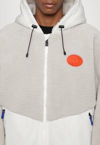 Felpa con cappuccio in grigio chiaro con un'esterno testurizzato, dettagli a coste bianche, zip frontale e una toppa arancione sul petto.