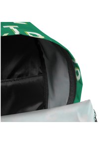 El interior de la bolsa verde presenta un forro negro y un compartimento con cremallera, con un patrón de texto en la tela exterior y un borde suave de color azul claro.