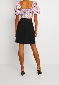 Rosa und gelb gemusterter Kurzarm-Top mit Puffärmeln, kombiniert mit schwarzen knielangen Shorts. Schwarze Absatzsandalen runden das Outfit ab.