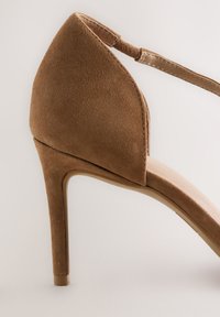 Rudos spalvos zomšinės aukštakulnės basutės, turinčios smailų priekį, čiurnos dirželį ir lygų kreminės spalvos pado paviršių. Kojinė plona, siaurėjanti aukštakulnio forma.