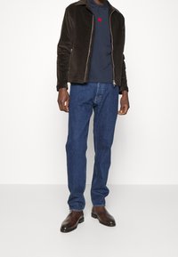 Ruskea cord-housutakki tummansinisellä poolopaidalla, jossa on punainen etiketti, yhdistettynä sinisiin farkkuihin ja tummanruskeisiin nahkabootseihin.