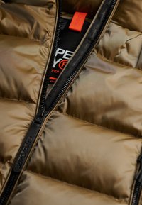 Zbliżenie częściowo rozpiętej brązowej kurtki puchowej pokazującej czarny zamek błyskawiczny z napisem „SUPERDRY” oraz czarną podszewkę z pomarańczowym i białym tekstem.