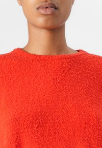 Pull en tricot texturé rouge avec un col rond. Le tissu semble doux, présentant une finition duveteuse et une couleur vibrante et uniforme sur toute sa surface.