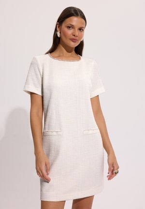 Kobieta w białej, teksturowanej sukience z krótkim rękawem, z perłową lamówką na kieszeniach i dekolcie, stojąca na tle jasnego, jednolitego tła.