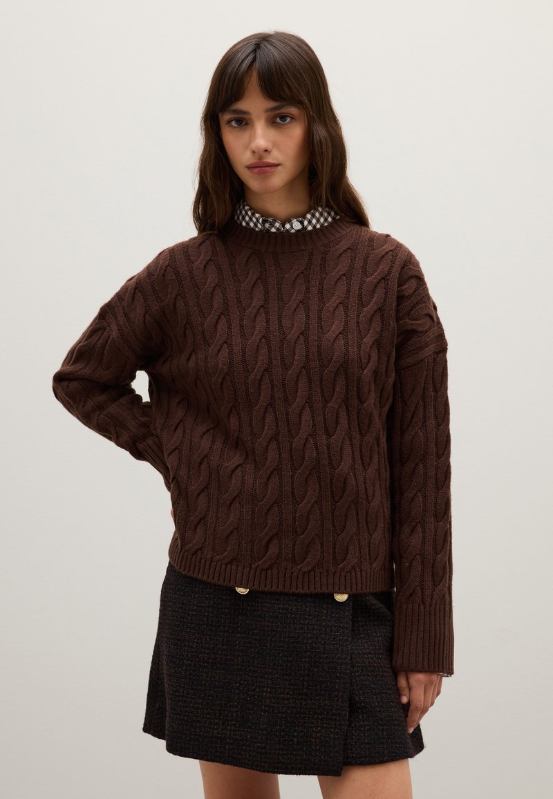 Bruin kabelgebreid sweater met geribde zoom, gedragen over een zwart-wit geblokt overhemd met kraag. Model poseert met de handen op de heupen.