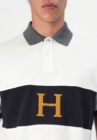 Paita polo, jossa on valkoinen tärkeä osa ja tummansininen vaaka-aita sekä brodeerattu kultainen "H"-logo, viimeistelty harmaalla kauluksella.