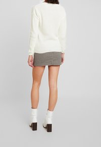 Kvinde iført hvid strikket sweater, sort-hvid ternet miniskørt og hvide hælede ankelstøvler, stående foran en ensfarvet grå baggrund.