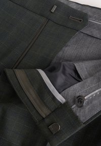 Close-up of dark suit jacket sleeve showing gray lining, button, and small metal clasp with visible stitching details.