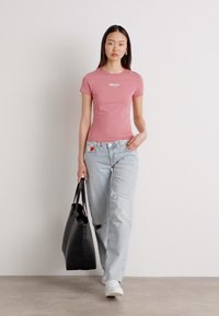 Růžové upnuté tričko s bílým logem, světle modré distressed džíny a černá kabelka. Modelka má na sobě bílé tenisky. Jednoduchý, ležérní outfit.