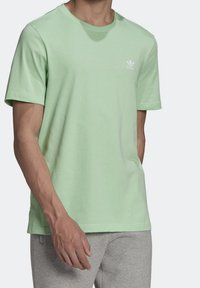 Mátově zelené bavlněné tričko s krátkým rukávem, kulatým výstřihem a bílým logem Adidas na levé straně hrudi. V páru se šedými tepláky.