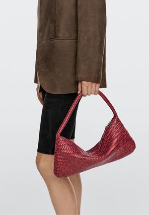 Person iført brun jakke og sort nederdel holder en rød vævet håndtaske med en enkelt skulderrem mod en ensfarvet baggrund.