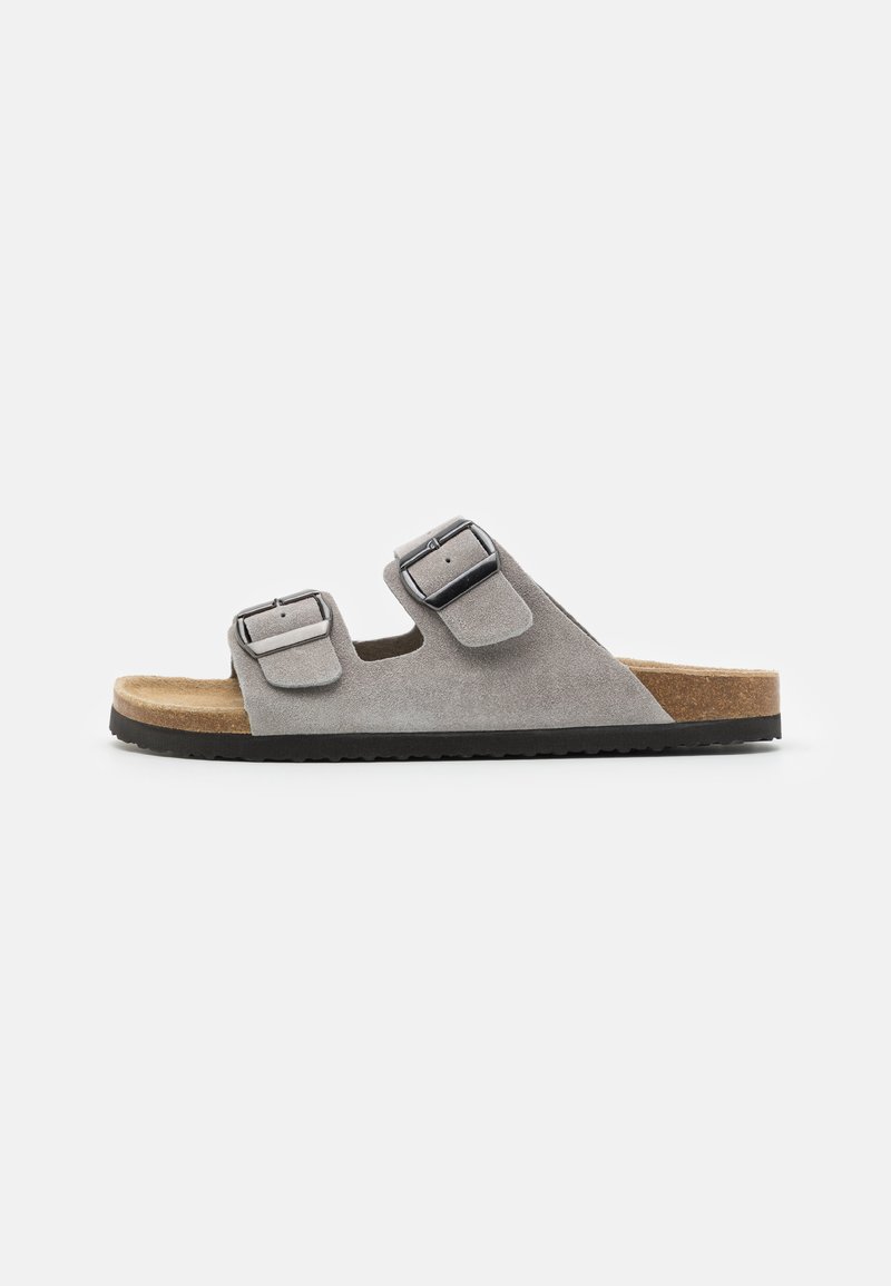 Sandal cinza em camurça, tipo slide, com duas fivelas ajustáveis, uma palmilha de cortiça e uma sola de borracha preta texturizada. Vista lateral, design minimalista.