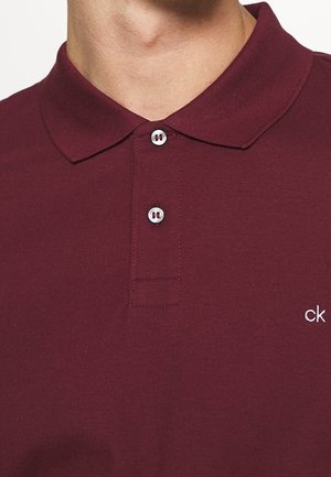 Mand iført en vinrød poloshirt med to hvide knapper og en lille hvid "ck"-logo broderet på venstre bryst.