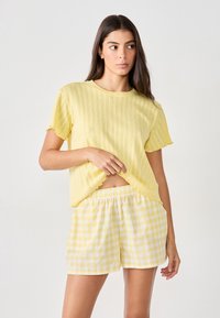 Top amarillo de manga corta con textura acanalada y bordes con volantes, combinado con pantalones cortos de cuadros con gingham en amarillo claro y blanco. Cintura elástica.