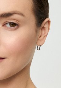 Srebrna kolczyki w kształcie obręczy, charakteryzujące się smukłym, otwartym wzornictwem z polerowanym wykończeniem, zawieszone na małżowinie usznej na neutralnym tle.