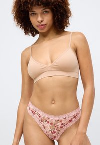 Μπεζ σουτιέν με λεπτές τιράντες, συνδυασμένο με ροζ δαντελωτό στρινγκ με περίπλοκες λεπτομέρειες και κυματιστές άκρες.