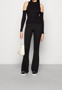 Czarny prążkowany top bez rękawów z odkrytymi ramionami, w zestawieniu z dopasowanymi czarnymi spodniami dzwonami. Trzyma małą białą torebkę.