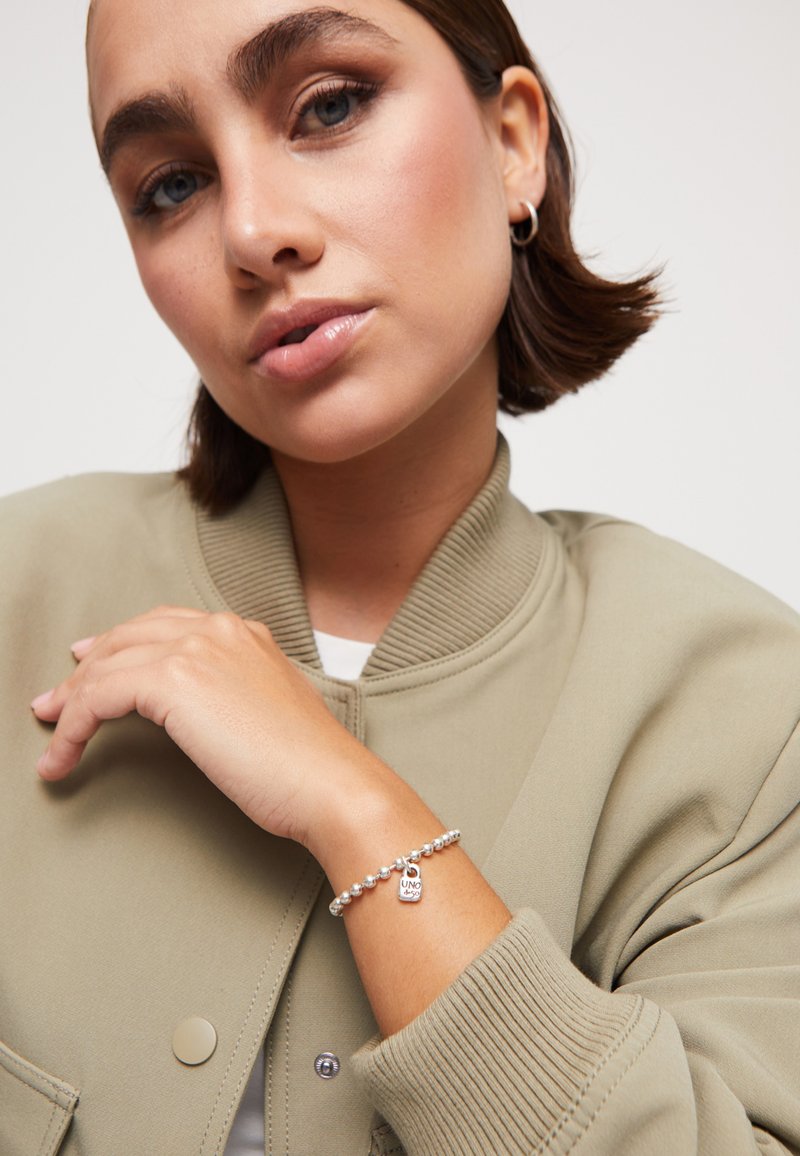 Pulsera de plata con cuentas y un charm, sobre una chaqueta bomber verde. Presenta una textura suave y bordes redondeados.