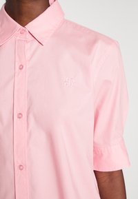 Kortärmad skjorta med krage i ljusrosa, tillverkad av ett mjukt tyg. Har knappstängning framtill och en diskret broderad logga på bröstet.