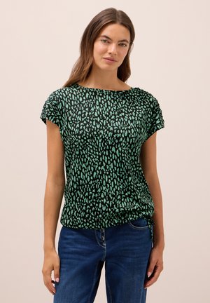 Femme avec de longs cheveux bruns portant un haut à manches courtes noir et vert à motifs et un jean bleu, se tenant contre un fond clair uni.