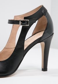 Chaussure à talon haut en cuir noir avec une bride à la cheville, détail de boucle argentée, côtés ouverts et finition lisse. Design à talon aiguille.