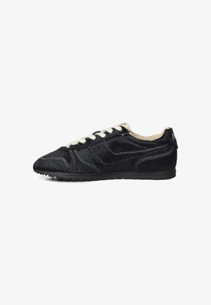 Zapatilla deportiva negra de corte bajo con cordones blancos, textura similar a piel, costuras blancas visibles y suela plana de goma sobre fondo blanco.