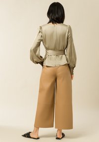 Satin olivgrüne Bluse mit Puffärmeln und gebundenem Saum, kombiniert mit weit geschnittenen braunen Hosen und schwarzen Rutschen-Sandalen.