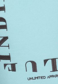 Tessuto di cotone azzurro chiaro con testo ricamato in marrone scuro. Il testo include "UNLIMITED APPAREL" in lettere maiuscole grassetto. Texture liscia.