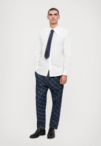 Camisa blanca de manga larga con cuello puntiagudo, combinada con pantalones marinos de patrón con un diseño geométrico y zapatos negros.