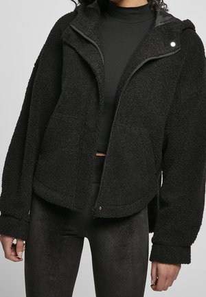 Femme portant une veste noire duveteuse zippée sur un crop top noir à col roulé et des leggings en velours noirs sur un fond uni.