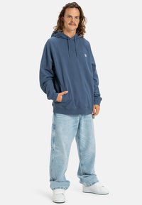 Sudadera azul marino hecha de una mezcla de algodón con un bolsillo delantero, capucha con cordones, puños acanalados, combinada con jeans de pierna ancha de color azul claro y zapatillas blancas.