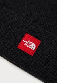 Berretto nero lavorato a maglia con un'etichetta rettangolare rossa recante testo bianco e un logo. Il tessuto appare morbido e texturizzato.