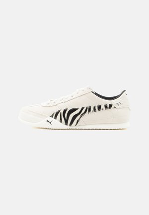 Baskets blanches Puma avec un logo latéral zébré noir et blanc, design bas et semelle blanche sur fond clair uni.