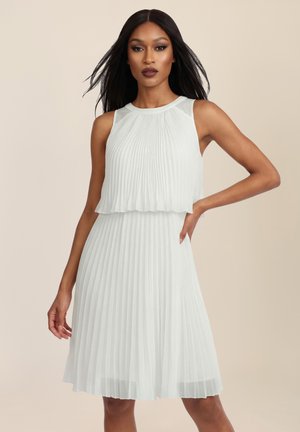 Frau mit langen schwarzen Haaren, die ein ärmelloses weißes, plissiertes Kleid trägt und mit einer Hand auf der Hüfte vor einem schlichten beigen Hintergrund posiert.