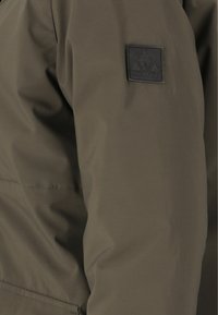 Olivgrön jacka med en slät, texturerad yta; har en svart fyrkantig logotyp på överarmen; designad för utomhusbruk.