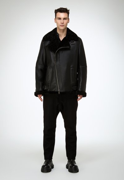 Schwarze Lederjacke mit einem breiten, flauschigen Kragen, Frontreißverschluss und dicker Fellbesatz am Saum. Modell trägt schwarze Hose und Schuhe.