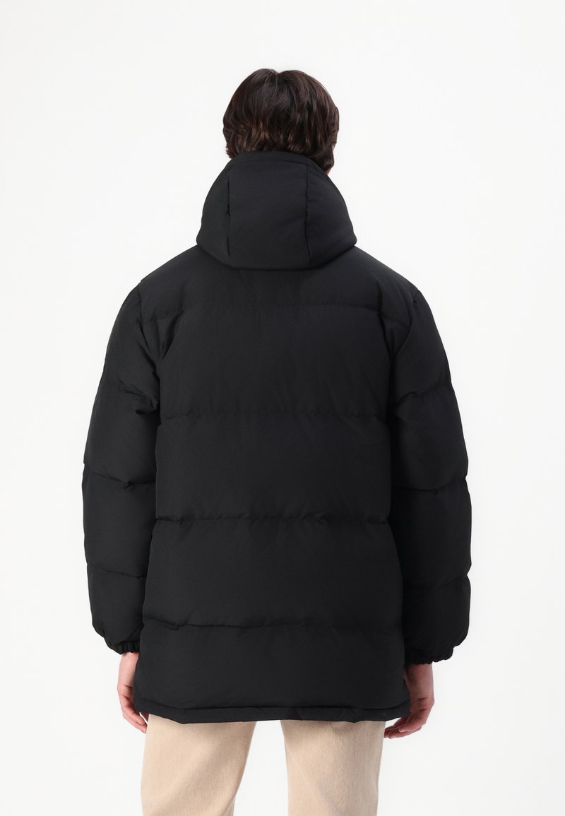Doudoune noire avec capuche, présentant des sections matelassées et une coupe ample. Le matériau semble lisse et fortement isolé, adapté aux temps froids.